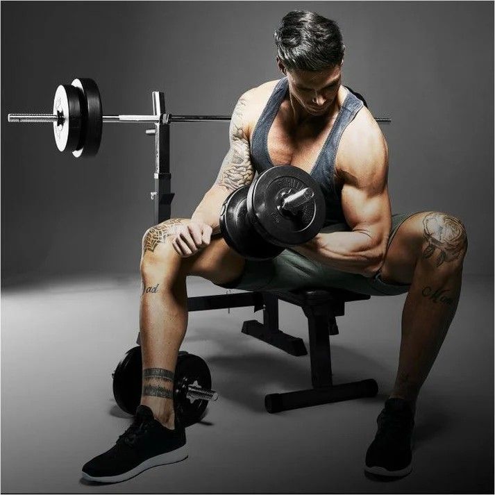 Muž cvičící biceps s jednoruční činkou na posilovací lavici se stojanem a osovou činkou v pozadí.