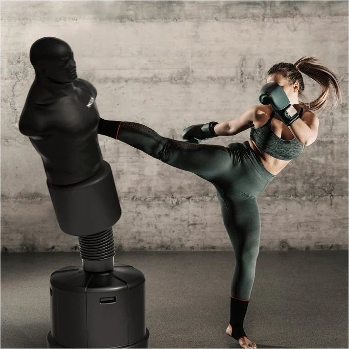 Žena v sportovním oblečení a rukavicích kopá do stojící boxovací figuríny s torzem na pevné základně ve cvičebním prostoru.